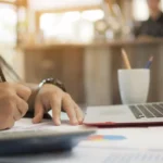 Person writes on paper at a desk with a laptop, mug, and papers in a bright workspace.