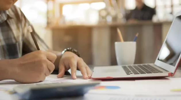 Person writes on paper at a desk with a laptop, mug, and papers in a bright workspace.