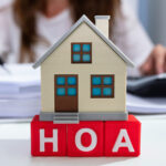 Miniature house on red blocks spelling HOA, with a person writing in the blurred background.