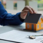 Person signs a contract at a desk with a small orange house model and keys nearby, indicating a real estate agreement
