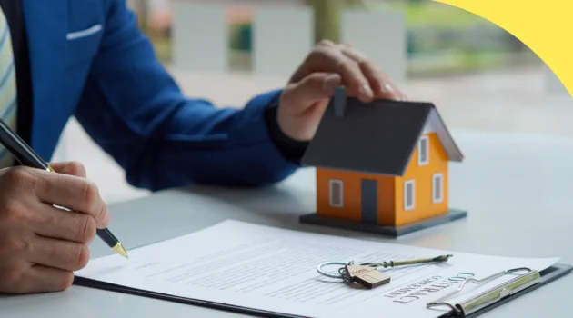 Person signs a contract at a desk with a small orange house model and keys nearby, indicating a real estate agreement